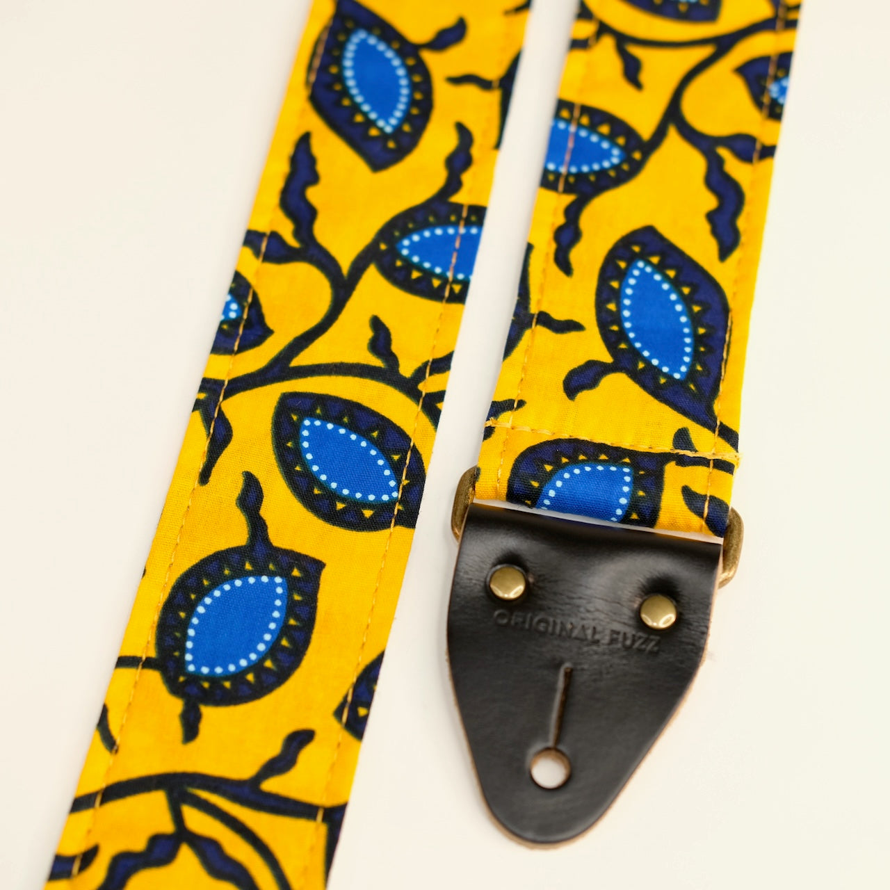 End-tab detail view of guitar strap in yellow and blue botanical African wax print fabric with black leather end-tab. Made in Nashville by Original Fuzz.