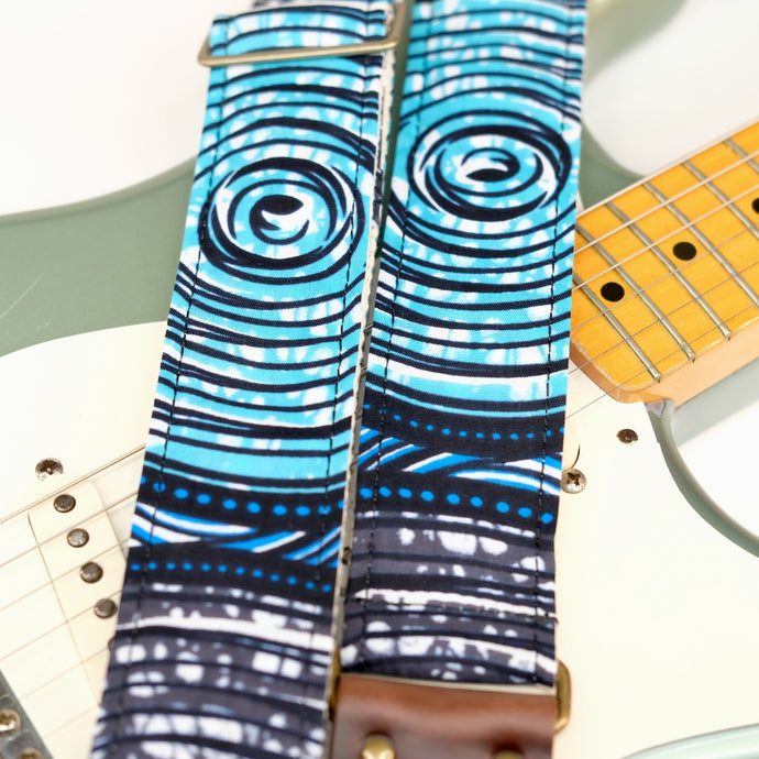 Wide view of guitar strap in white, black, blue, teal, and gray swirled African wax print fabric with brown leather end-tab. Made in Nashville by Original Fuzz.