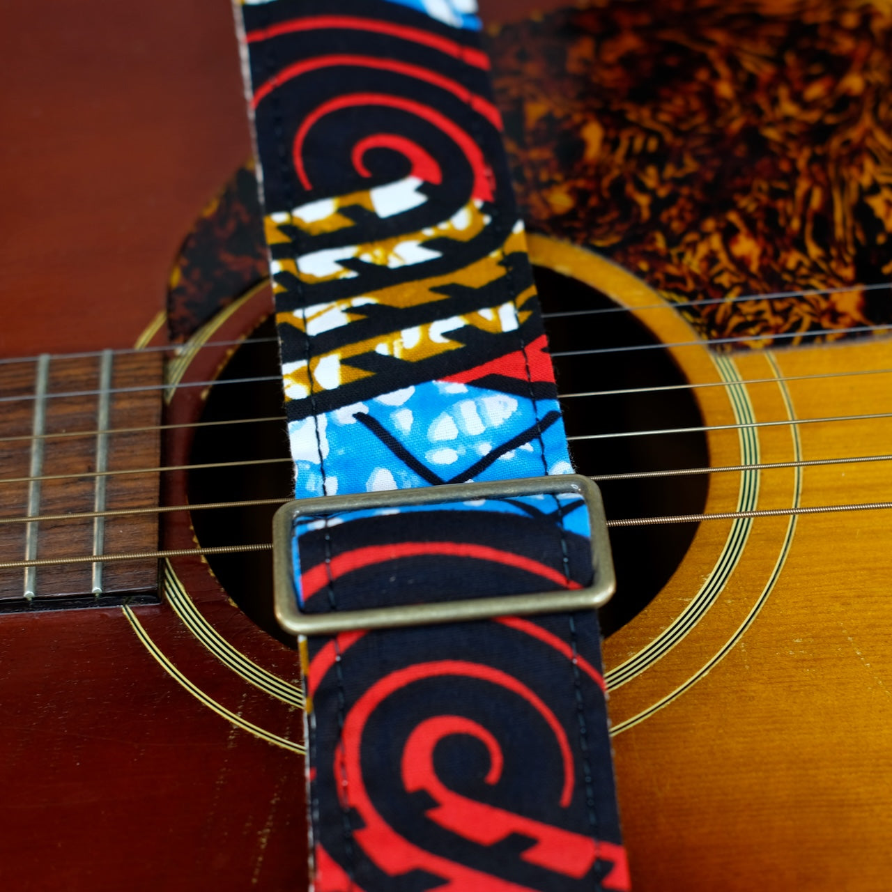 Buckle detail of guitar strap in black, red, blue, and tan swirled African wax print fabric with black leather end-tab. Made in Nashville by Original Fuzz.