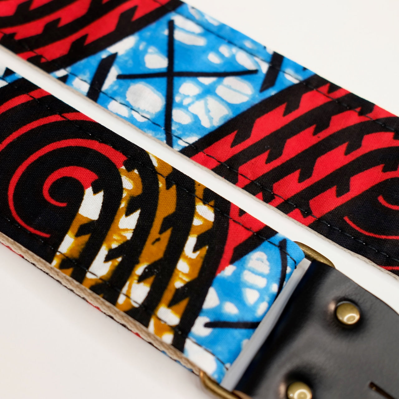 Fabric detail view of guitar strap in black, red, blue, and tan swirled African wax print fabric with black leather end-tab. Made in Nashville by Original Fuzz.