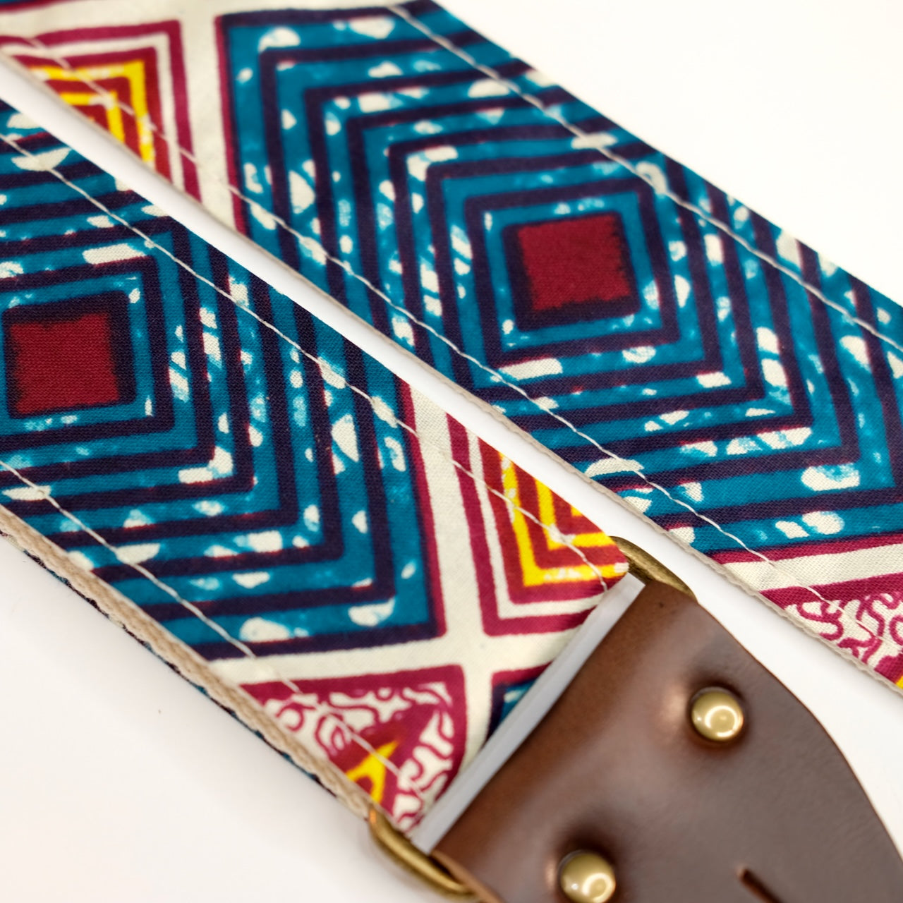 Fabric detail view of guitar strap in dark teal, burgundy, cream, and yellow diamond tile African wax print fabric with brown leather end-tab. Made in Nashville by Original Fuzz.