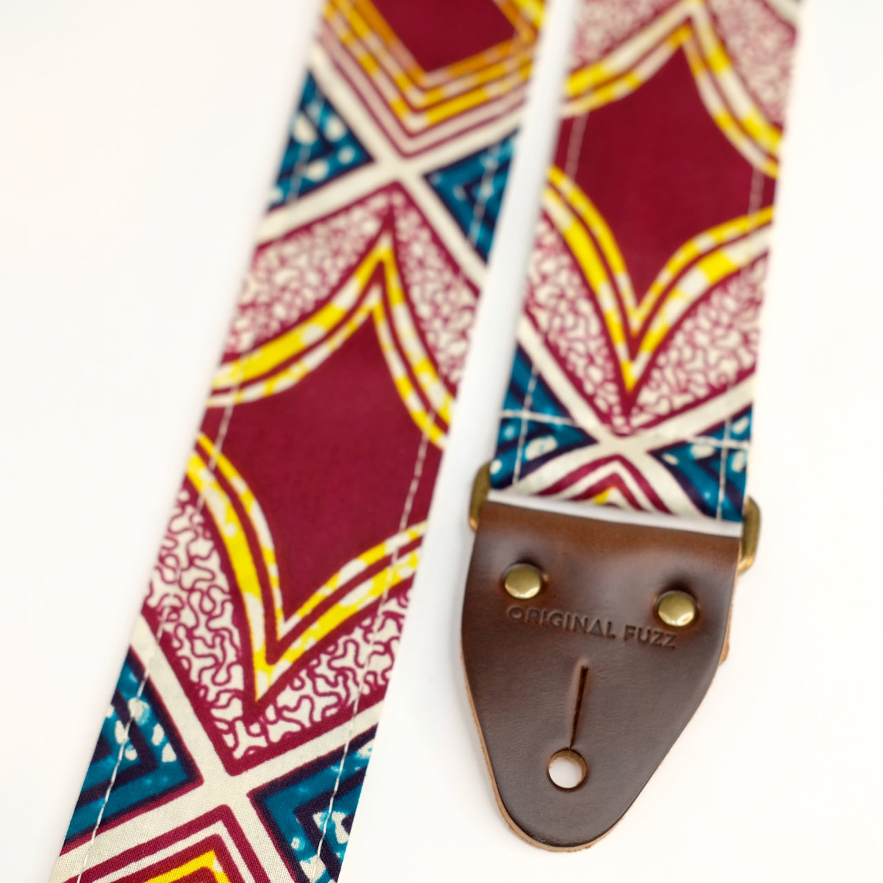 End-tab detail view of guitar strap in burgundy, cream, yellow, and dark teal diamond tile African wax print fabric with brown leather end-tab. Made in Nashville by Original Fuzz.