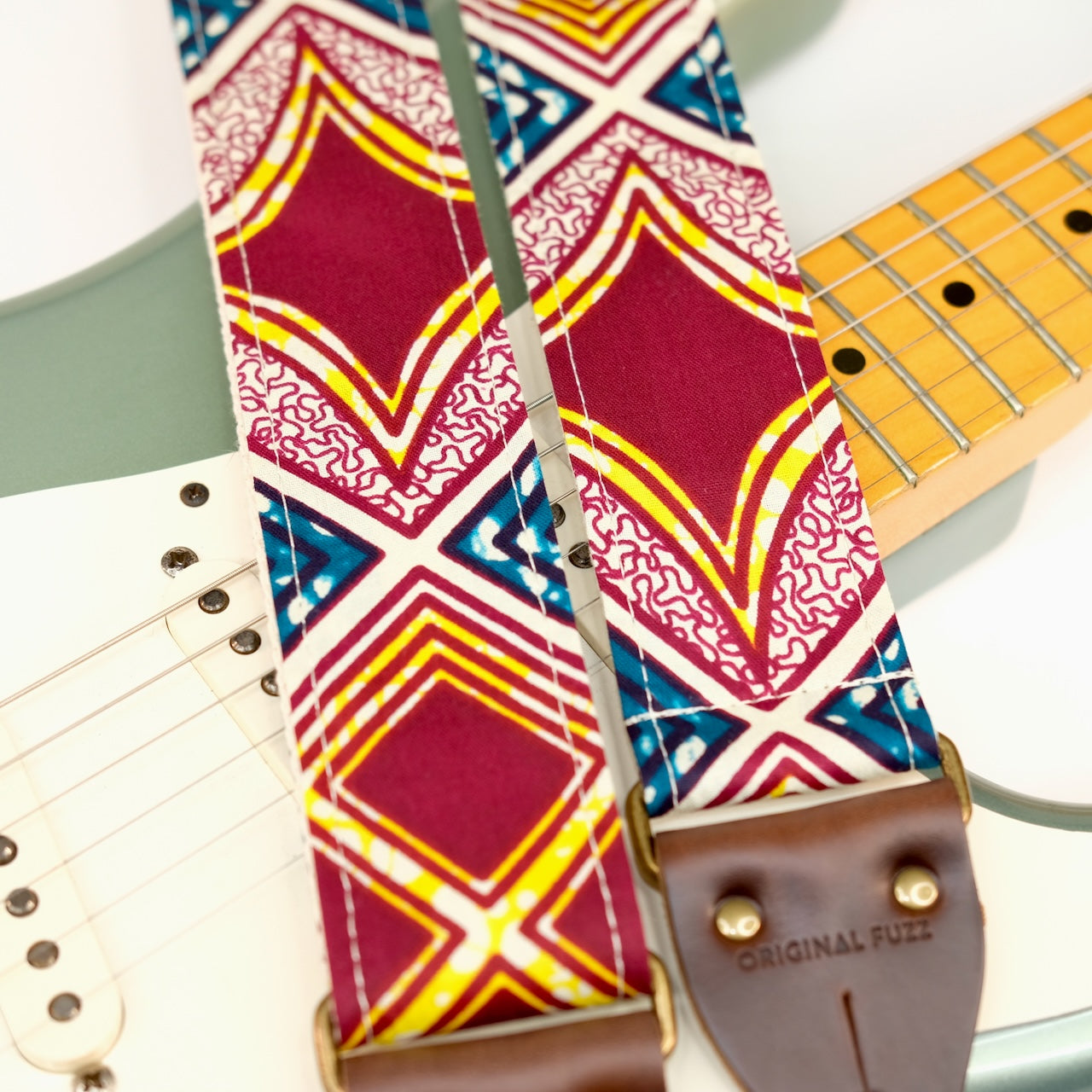 Wide view of guitar strap in burgundy, cream, yellow, and dark teal diamond tile African wax print fabric with brown leather end-tab. Made in Nashville by Original Fuzz.