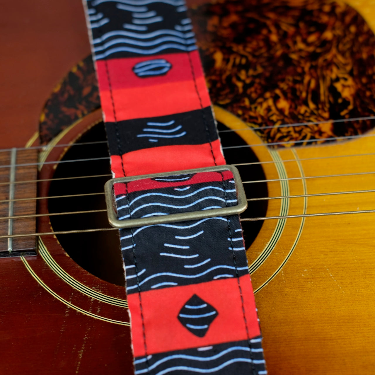 Buckle detail of guitar strap in black, red, and white striped African wax print fabric with black leather end-tab. Made in Nashville by Original Fuzz.