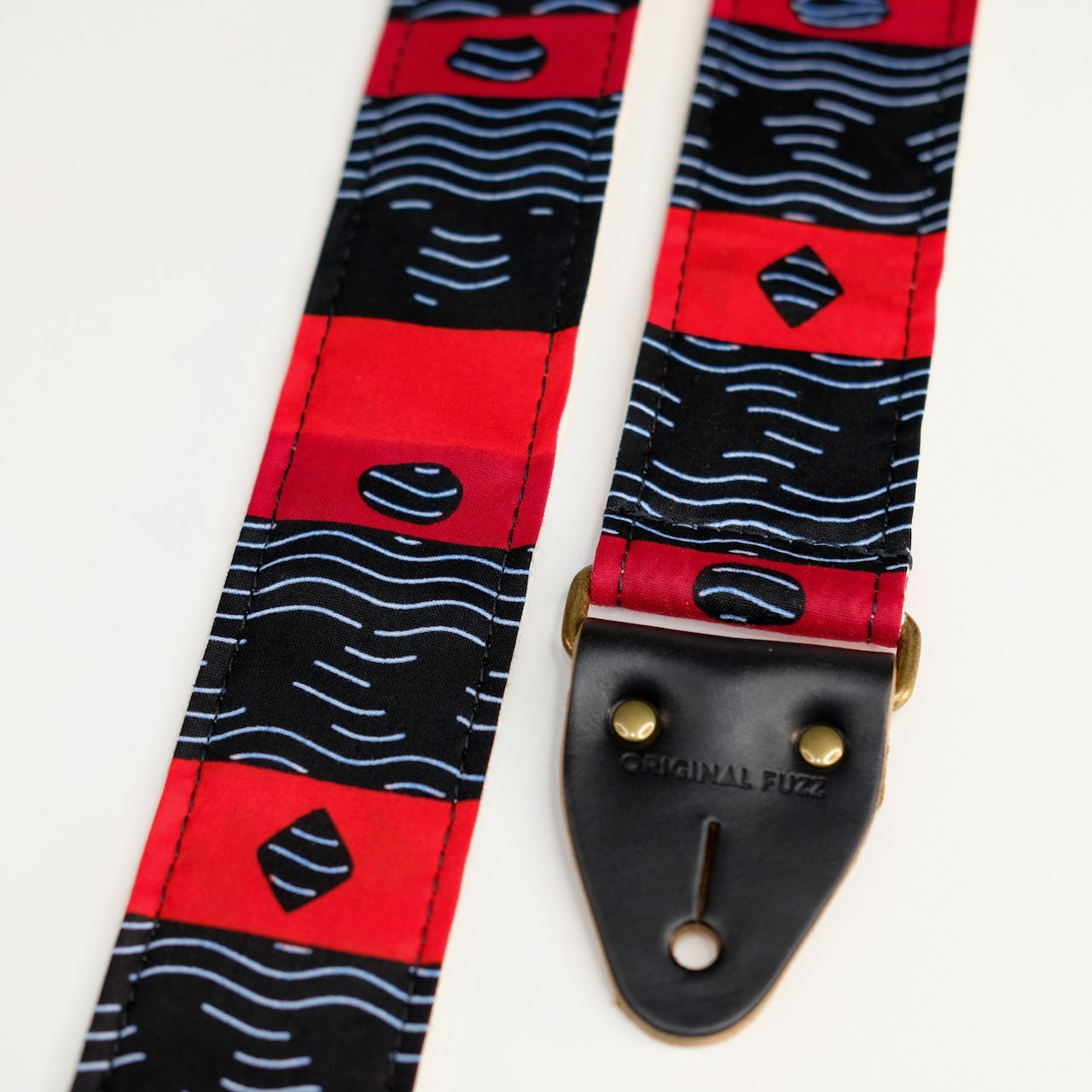 End-tab detail view of guitar strap in black, red, and white striped African wax print fabric with black leather end-tab. Made in Nashville by Original Fuzz.